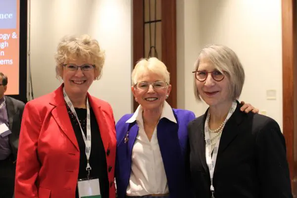 image: three women in business attire with light skin and light hair in a conference setting holding each other and smiling at the camera