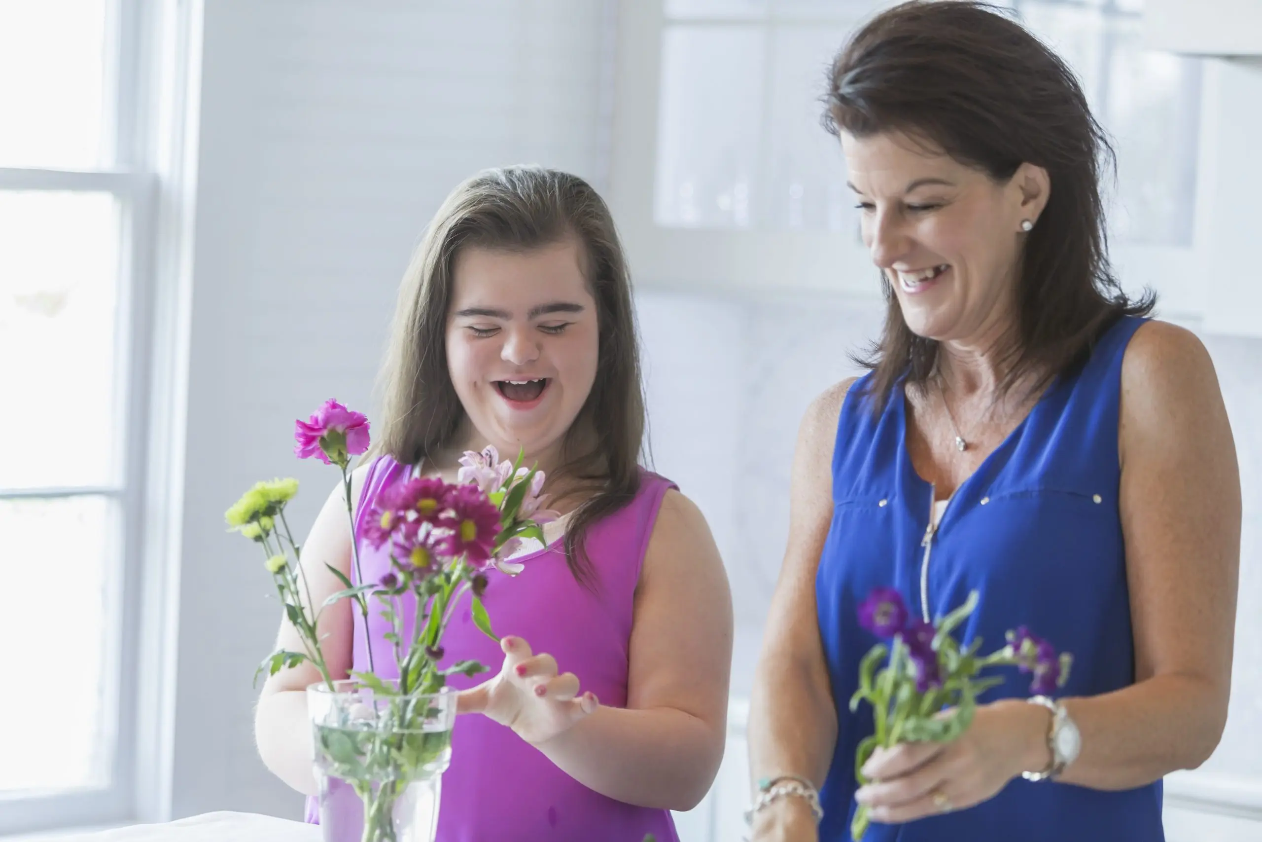 image: a woman and a girl with light skin and dark hair in casual attire, the girl with obvious disabilities smiling at some flowers