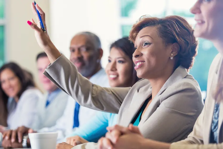 image of a woman with dark hair and skin in the middle of multiple diverse people raising her hand in a meeting office setting